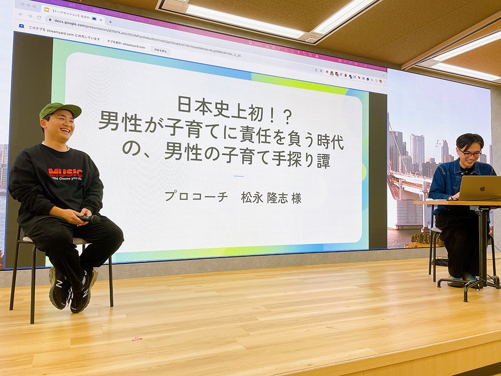 スピーカーの松永様、MCの大串がステージ上で対談している写真。スクリーンには、「日本史上初！？男性が子育てに責任を負う時代の、男性の子育て手探り譚」というセッションタイトルが映し出されている。