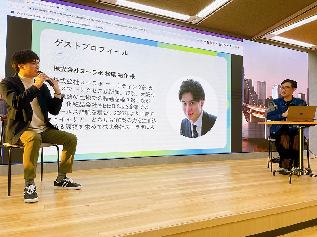 スピーカー松尾様とMCの大串がステージ上で対談している写真。スクリーンには、松尾様のプロフィールが映し出されている。